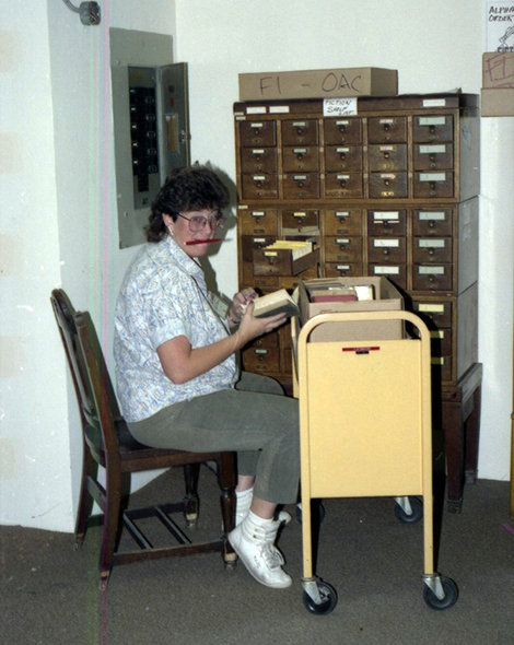 women seated with book cart in front of her and a pencil in her mouth