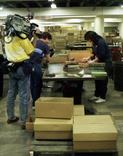 man with a camera filming 2 people cleaning books