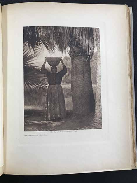 a Native American woman photographed holding a basket on her head among palm trees