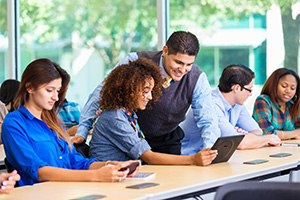 class of adults with teacher showing student something on laptop