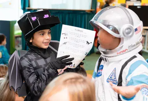 Two children, one in a bat costume and one in a spacesuit costume, talk to each other at a library event.