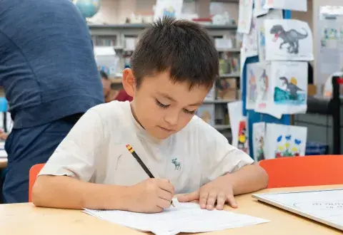 A young boy sits at a table in a library and writes on a piece of paper with a pencil