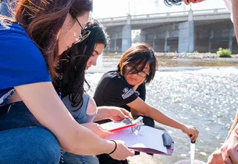 3 individuals with clipboard and note-taking materials squat near river