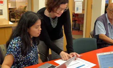 Multiple adults sitting at or standing near a desk, two with laptops, one with a sheet of paper