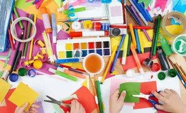 An assortment of craft supplies, paints, markers and hands with scissors cutting paper.