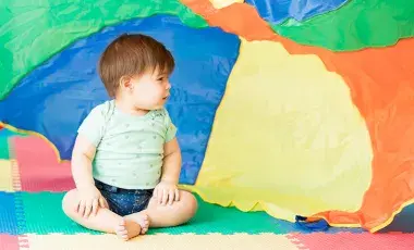 A baby sits on a colorful mat, with a colorful cloth waving behind them.