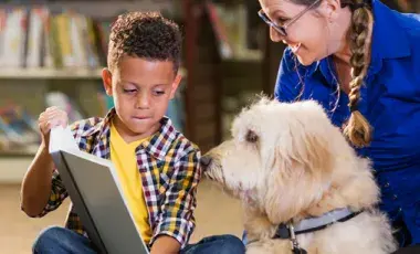 A young boy sitting in a library holds open a book for a white, shaggy dog while an encouraging woman looks on. 