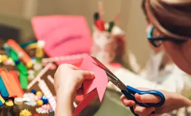 A woman cuts pink paper, art supplies are on a table in front of her.
