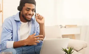 a young African-American man smiling sitting on a couch, looking down at a laptop that's on a small table, with headphones on