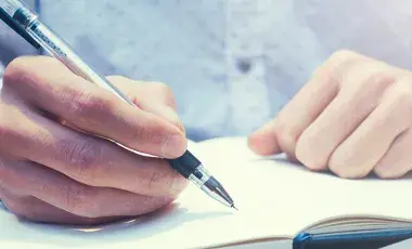 Close-up of two hands resting on an open journal, with the right hand holding a pen to write.