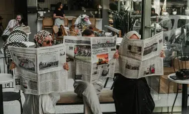 Three ladies sitting in chairs in a row holding up newspapers in front of their faces