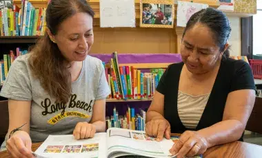 Two women sitting at a table reading a book.