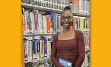 a tall woman wearing black glasses and holding a Dr. Seuss book standing next to tall library bookshelves