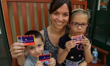 A woman and two children sit on a wooden bench, holding their Los Angeles Public Library Cards and smiling at the camera.