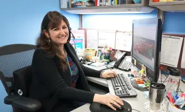 A librarian sits at a desk in an office. She is wearing a phone headset and smiling at the viewer. 