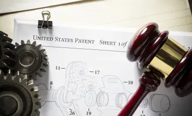 A wooden gavel and metal gears rest on top of a patent application with technical drawings.