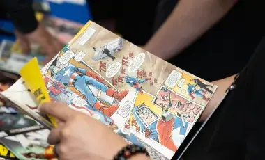 A close up of a graphic novel with colorful panes being held by a hand wearing a bracelet.