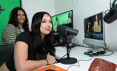 Two young women sit in a recording studio, with the foremost one seated at a microphone with computers and equipment in the background.
