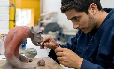 A man sculpts a fanciful creature from clay using pottery tools.