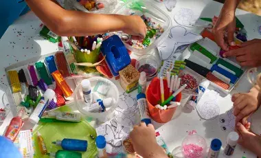 An assortment of craft supplies, paints, markers and hands with scissors cutting paper.