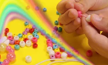 Hands making colorful friendship bracelets with rainbow beads on a bright yellow background.