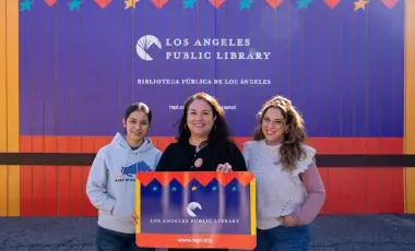 2 librarians and a clerk holding a giant library card stand in front of the temporary building