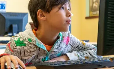 Boy sitting at computer looking at computer screen with hand on mouse
