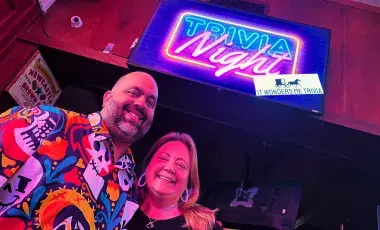 The hosts of It Wonders Me Trivia next to a picture of their logo of a covered wagon silhouette 