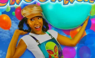 A smiling woman wearing a cap holds up a blue beach ball against a backdrop of balloons