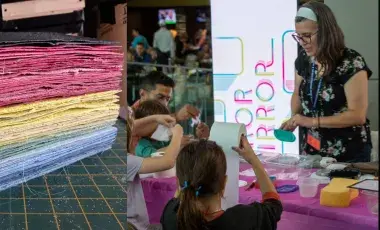 A tall stack of colorful, textured handmade paper on the left and a busy community papermaking workshop on the right