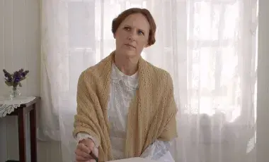 Molly Shannon dressed as Emily Dickinson sits in a sunlight room holding a pen