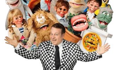Smiling puppeter is surrounded by various puppets. 