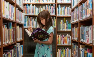 Head & shoulder view of brunette, white girl, reading book her in arms, surrounded by book-filled shelves