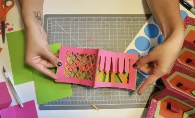 hands displaying pop up book art project, scissors and paper on table