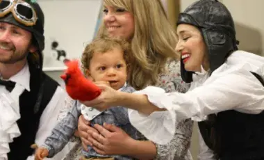 Two LA Opera performers in costume holding a bird prop in front of a young mother with her baby toddler.