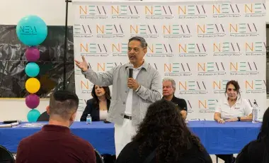 A man holding a microphone gestures in front of an audience with seated panelists behind him.