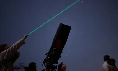 A person points to the night sky using a laser while standing next to a telescope. 