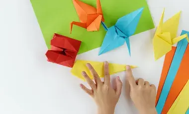 A pair of hands are folding some colored paper to make paper cranes for Origami. 
