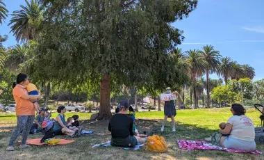 librarian reads a picturebook to a group of adult caregivers and toddlers on a grass lawn under a shady tree