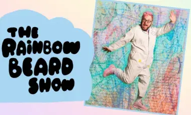 A photo of Ricky Rainbow Beard, with pink hair, a jumpsuit, and pink shoes, dancing in front of a colorful background that looks like crayon scribbles