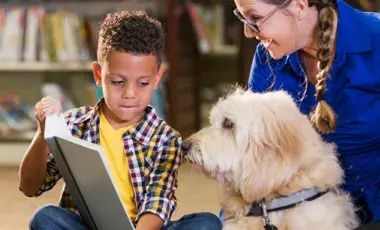 A young boy reads to a white dog while a smiling woman watches