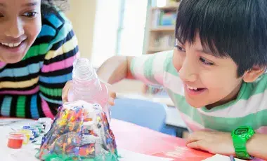 Two smiling children bent over a volcano science experiment pouring in the solution and waiting for it to bubble over.