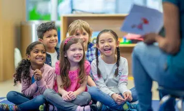 A group of children are smiling and laughing while listening to a Storyteller read a book. 