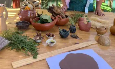 Different shaped clay jars holding corn, jicama and other plants sitting on top of a table. A flattened piece of clay is to the side.