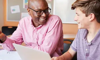 An adult male and a teen male sitting at a table with open notebooks and a laptop. They appear to be discussing academics.