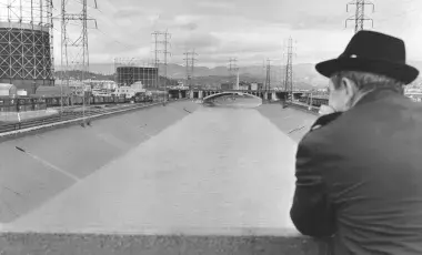 Person in overcoat and wide-brimmed hat facing away from the camera, looking down at the Los Angeles River with industrial tanks in the background.