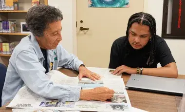 A person helping another person to read the newspaper. 