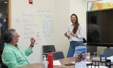 A teacher standing in front of a white board full of words. A student rising is their hand.