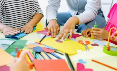 Hands are around a table creating art out of colorful materials. 