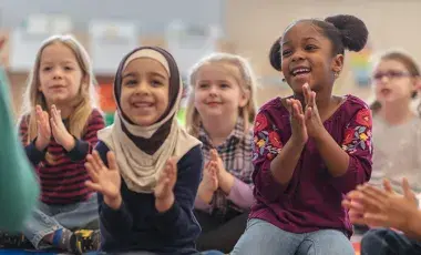 A group of children are smiling and laughing while listening to a Storyteller read a book. 